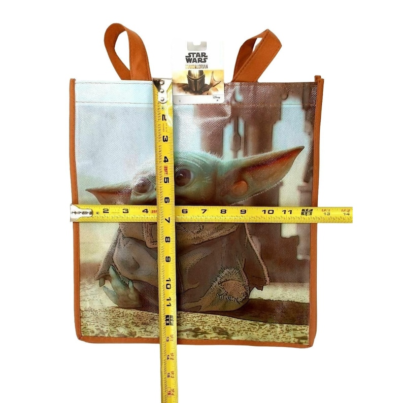 Op de afbeelding: Een herbruikbare boodschappentas met een kleurrijk beeld van "The Child" uit de Star Wars-serie "The Mandalorian". De tas is gemaakt van geweven stof en heeft oranje handvatten. De tas is ongeveer 28 cm breed en 36 cm hoog.