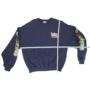 May include: Navy blue sweatshirt with the Ron Jon Surf Shop logo on the chest and floral and parrot graphics on the sleeves. The sweatshirt has long sleeves and a crew neck. The overall length is approximately 28 inches.