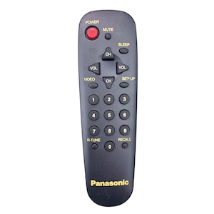 May include: A black Panasonic remote control with a red power button. The remote has buttons for volume, channel, and numbers 0-9. The text "Panasonic" is printed in gold at the bottom.