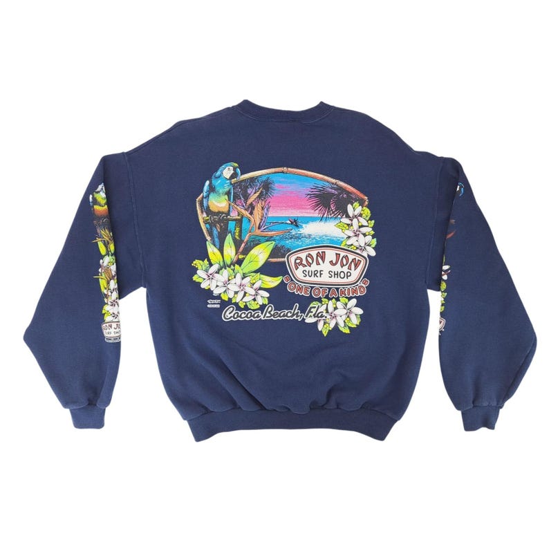 May include: Navy blue sweatshirt with a colorful graphic featuring a parrot, palm trees, and ocean scene. The text "Ron Jon Surf Shop Cocoa Beach, Fla." is printed on the back. Floral and leaf designs are on the sleeves.