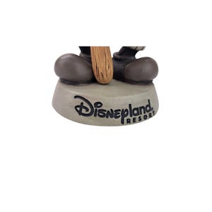 Peut inclure: Base de figurine grise claire avec le logo Disneyland Resort en noir. Les pieds de la figurine et une partie d'un objet en bois sont visibles.