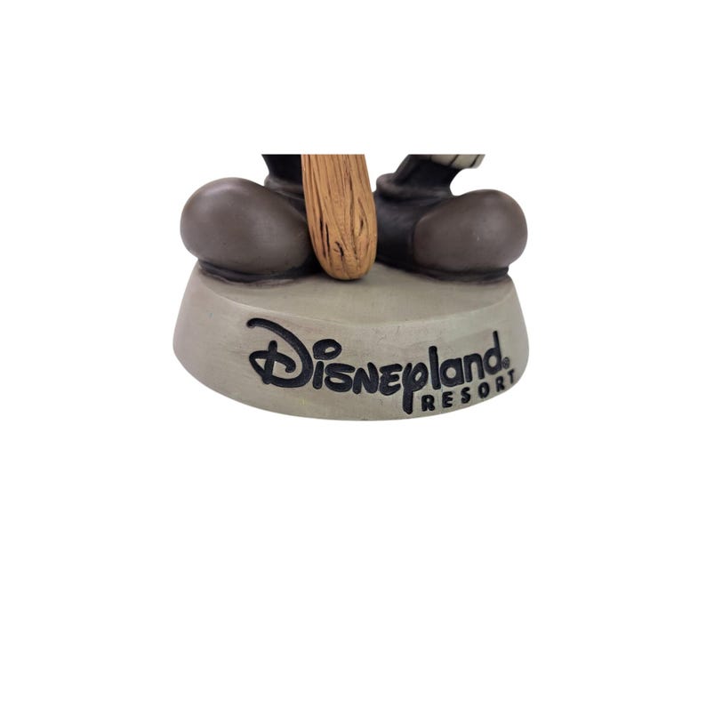Peut inclure: Base de figurine grise claire avec le logo Disneyland Resort en noir. Les pieds de la figurine et une partie d'un objet en bois sont visibles.