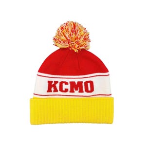 Può includere: Un berretto lavorato a maglia rosso, bianco e giallo con un pompon. Il cappello presenta le lettere "KCMO" in rosso su una fascia bianca. Il bordo è giallo.