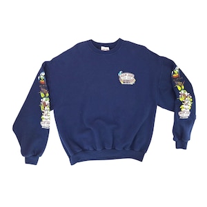 May include: Navy blue sweatshirt with the text "RON JON SURF SHOP" on the chest and sleeves. The sleeves feature a colorful floral and bird design. The sweatshirt is made of a soft material.
