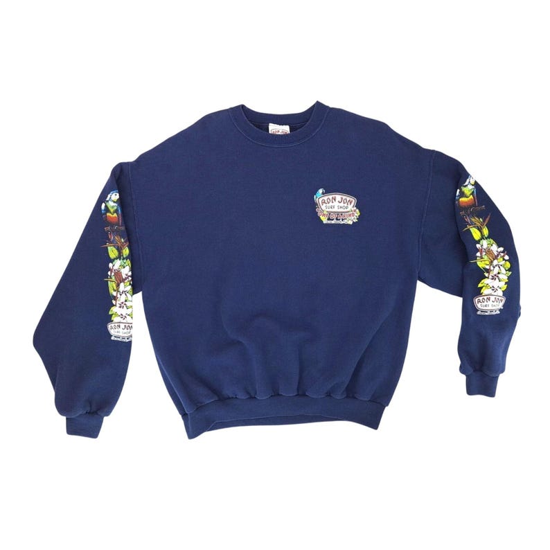 May include: Navy blue sweatshirt with the text "RON JON SURF SHOP" on the chest and sleeves. The sleeves feature a colorful floral and bird design. The sweatshirt is made of a soft material.