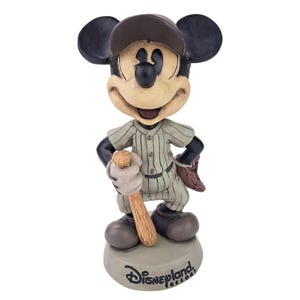 Peut inclure: Figurine de Mickey Mouse en uniforme de baseball, tenant une batte et portant une casquette. La figurine est dans une pose classique, avec un gant de baseball sur le dos. La base est grise et porte le logo Disneyland Resort.