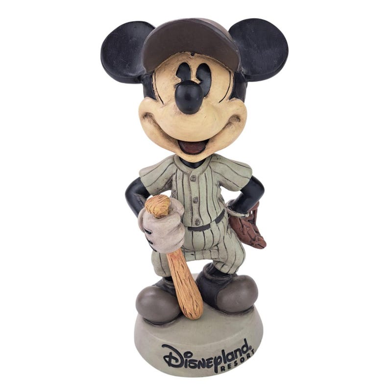 Peut inclure: Figurine de Mickey Mouse en uniforme de baseball, tenant une batte et portant une casquette. La figurine est dans une pose classique, avec un gant de baseball sur le dos. La base est grise et porte le logo Disneyland Resort.