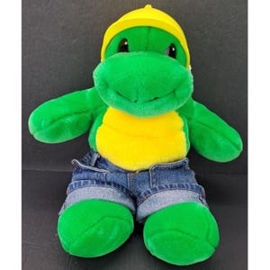 May include: A plush turtle toy with bright green fur, a yellow belly, and a yellow cap. The turtle is wearing blue denim shorts with cuffed hems. The toy features large black eyes and a soft, rounded snout, presented in a seated position.