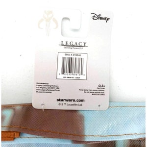 Op de afbeelding: Een witte en blauwe herbruikbare boodschappentas met een Star Wars-thema en een Mandalorian-personageontwerp. De tas heeft een wit label met de tekst "LEGACY Licensing Partners, LP" en "SKU # 315046". Het label bevat ook een barcode en de tekst "starwars.com &copy; & &trade; Lucasfilm Ltd."