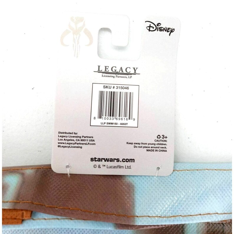 Op de afbeelding: Een witte en blauwe herbruikbare boodschappentas met een Star Wars-thema en een Mandalorian-personageontwerp. De tas heeft een wit label met de tekst "LEGACY Licensing Partners, LP" en "SKU # 315046". Het label bevat ook een barcode en de tekst "starwars.com &copy; & &trade; Lucasfilm Ltd."