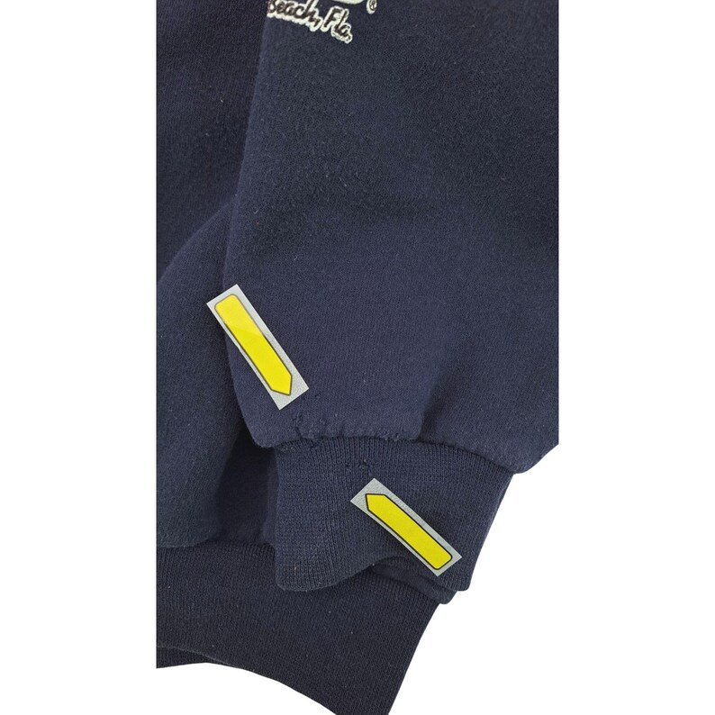 May include: Navy blue sweatshirt with yellow arrow-shaped accents on the sleeves. The cuffs are ribbed. The text "Beach, Fla." is visible on the upper left side of the sweatshirt.