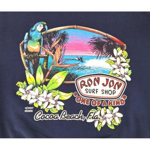 May include: Navy blue t-shirt featuring a tropical scene with a parrot, palm trees, and a surfer. The design includes the text "Ron Jon Surf Shop" and "Cocoa Beach, Fla.", surrounded by flowers.