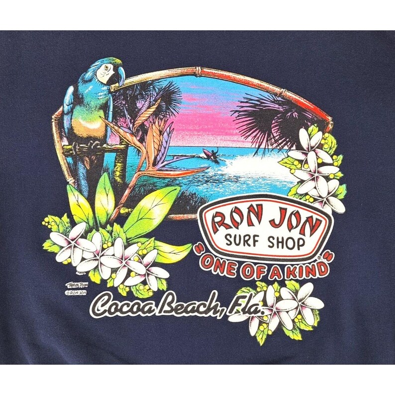 May include: Navy blue t-shirt featuring a tropical scene with a parrot, palm trees, and a surfer. The design includes the text "Ron Jon Surf Shop" and "Cocoa Beach, Fla.", surrounded by flowers.