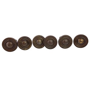 Puede incluir: Un juego de seis pomos circulares de metal para armarios con un acabado de bronce oscuro desgastado. Cada pomo presenta una cúpula central elevada rodeada por un diseño de anillos concéntricos, mostrando una apariencia texturizada.