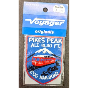 May include: A rectangular embroidered patch featuring a red cog railroad train on a blue background with a white mountain peak. The text reads Pikes Peak Alt 14,110 Ft. Cog Railroad. It is attached to a blue Voyager Originals header card.