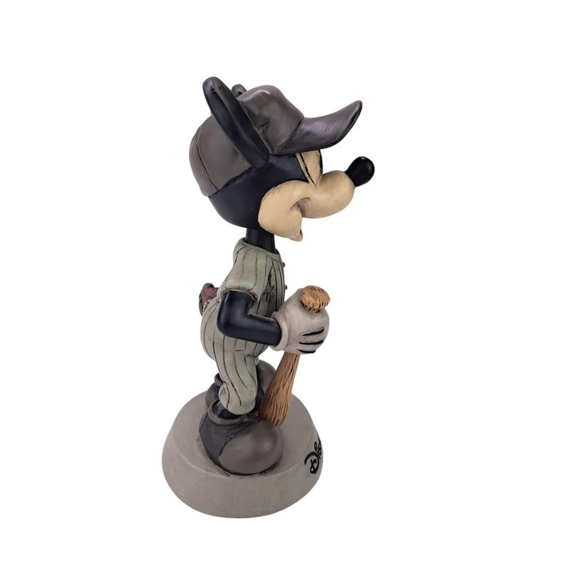 Peut inclure: Figurine d'un personnage de dessin anim&eacute; en uniforme de baseball, tenant une batte. La figurine est principalement noire, avec un visage beige et une casquette grise. L'uniforme est vert clair avec des rayures verticales. La figurine est sur une base grise.