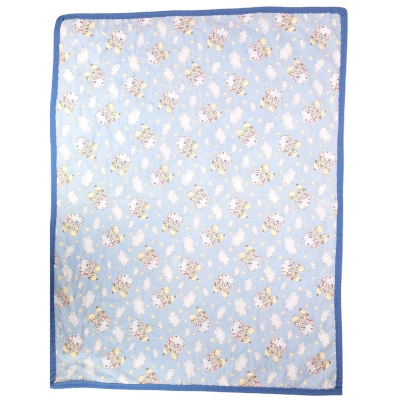 May include: A baby blanket featuring a light blue backdrop adorned with a repeating pattern of cartoon sheep, clouds, and stars. The blanket has a blue border and is likely made of soft fabric.