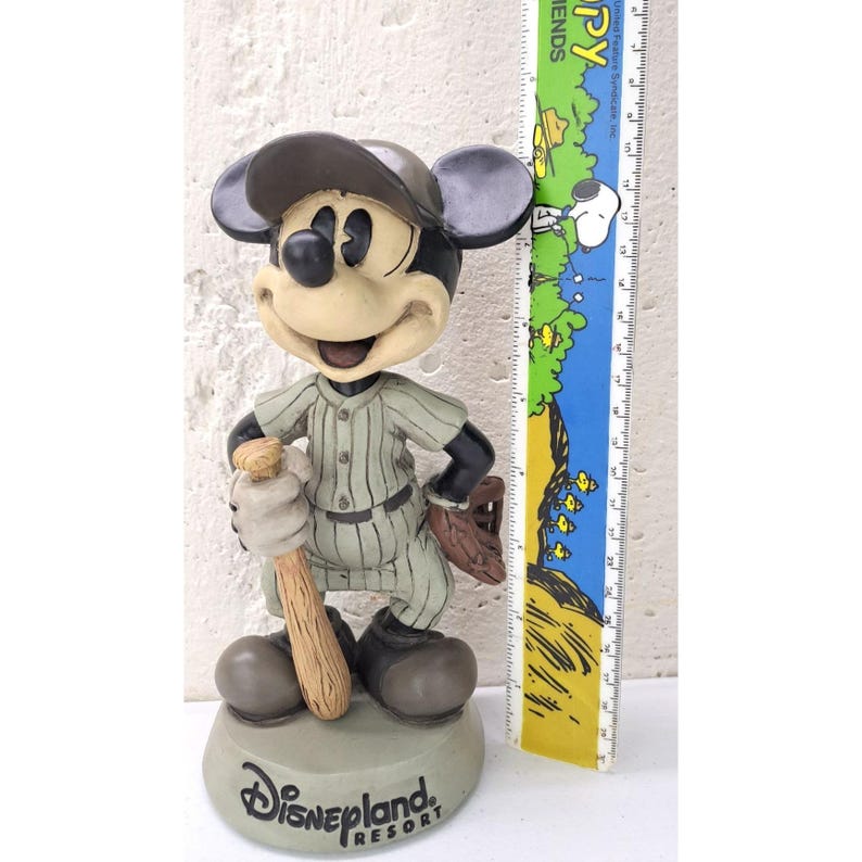 Peut inclure: Figurine de Mickey Mouse de Disneyland Resort en uniforme de baseball, tenant une batte et portant une casquette. La figurine est grise, noire et beige, avec le logo Disneyland Resort sur la base.