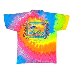 Puede incluir: Una camiseta te&ntilde;ida con un remolino vibrante de colores que incluyen azul, rosa, naranja y verde. La parte posterior de la camiseta presenta un gr&aacute;fico con las palabras "LESS STRESS" y "MORE FUN" alrededor de un caim&aacute;n de dibujos animados relaj&aacute;ndose en una playa. Tambi&eacute;n se ve el texto "RON JON".