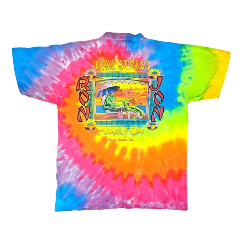 Puede incluir: Una camiseta te&ntilde;ida con un remolino vibrante de colores que incluyen azul, rosa, naranja y verde. La parte posterior de la camiseta presenta un gr&aacute;fico con las palabras "LESS STRESS" y "MORE FUN" alrededor de un caim&aacute;n de dibujos animados relaj&aacute;ndose en una playa. Tambi&eacute;n se ve el texto "RON JON".