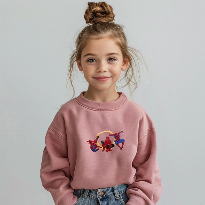 May include: A pink sweatshirt with an embroidered Spider-Man design. The design shows Spider-Man in action. The sweatshirt is worn by a young person with a bun hairstyle and blue jeans.