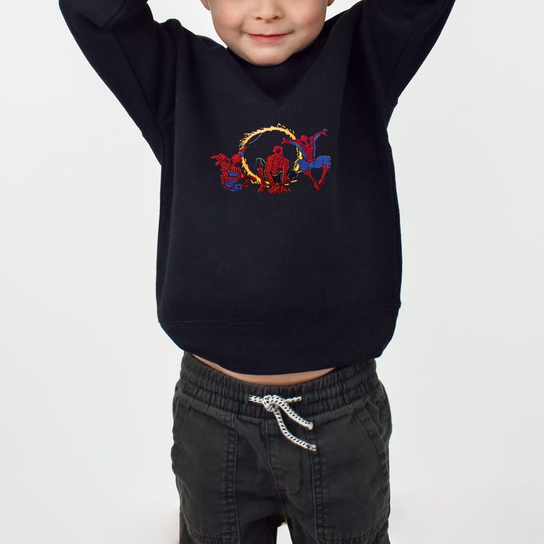 May include: Black crew neck sweatshirt with an embroidered design of three superheroes within a yellow ring. The superheroes are in red, blue, and yellow. The sweatshirt is paired with dark grey trousers.