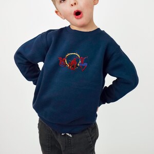 May include: Navy blue sweatshirt with an embroidered Spider-Man design. The design features Spider-Man in action poses with a yellow and orange web-like circle. The sweatshirt is made of a soft material.