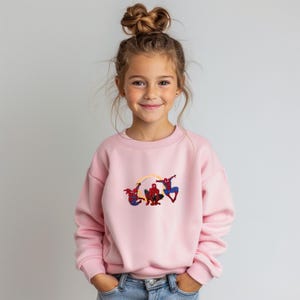 May include: A pale pink sweatshirt with an embroidered design of three Spider-Man figures. The figures are in red, blue, and black, with yellow web lines. The sweatshirt is worn by a young girl with her hair up in a bun.