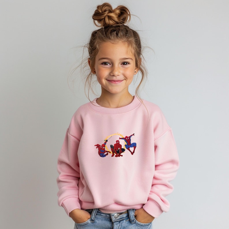 May include: A pale pink sweatshirt with an embroidered design of three Spider-Man figures. The figures are in red, blue, and black, with yellow web lines. The sweatshirt is worn by a young girl with her hair up in a bun.