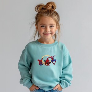 May include: A pale blue sweatshirt with an embroidered design of Spider-Man. The superhero is shown in red and blue, with a yellow web effect. The sweatshirt is a casual style, suitable for everyday wear.