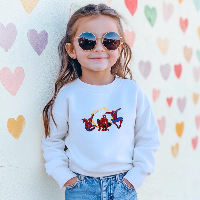 May include: A child's white sweatshirt featuring an embroidered design of three Spider-Man characters. The sweatshirt is paired with blue jeans. The girl is wearing round sunglasses. The background is a wall decorated with heart shapes.