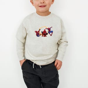 May include: A light grey sweatshirt with an embroidered design of three Spider-Man figures. The figures are in red, blue, and black, with yellow web details. The sweatshirt has long sleeves and a crew neck.
