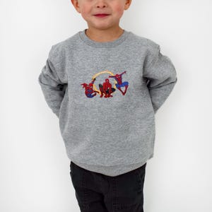 May include: A grey crew neck sweatshirt with an embroidered design of Spider-Man figures. The design features three Spider-Man characters in red and blue, with yellow accents. The sweatshirt is a casual style.