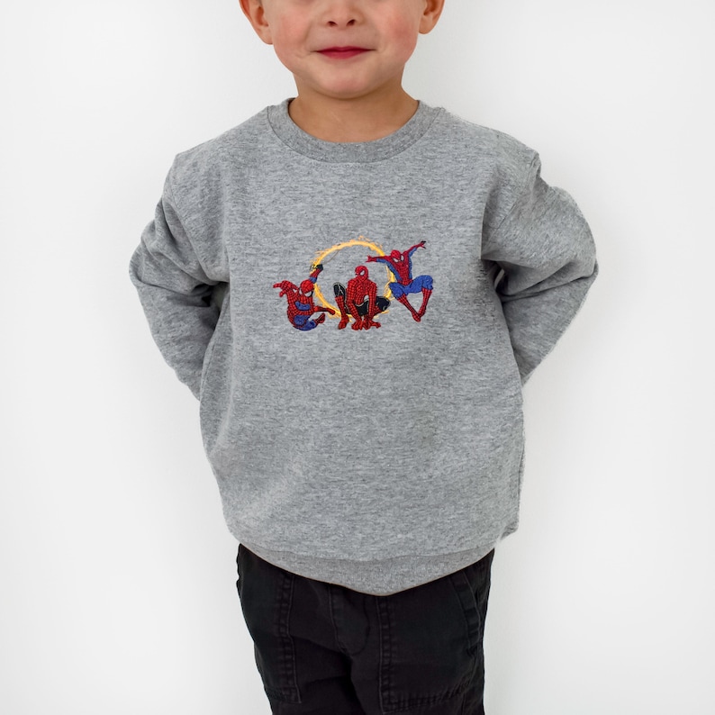 May include: A grey crew neck sweatshirt with an embroidered design of Spider-Man figures. The design features three Spider-Man characters in red and blue, with yellow accents. The sweatshirt is a casual style.