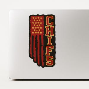 May include: A sticker featuring the word "CHIEFS" in gold, stacked vertically. The left side of the sticker is a red, white, and gold design resembling a flag. The top left corner has gold stars.