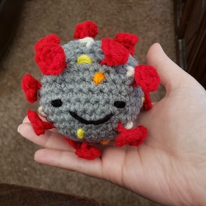May include: A gray crocheted plush toy shaped like a virus with red, yellow, and white accents. The toy has a smiling face.