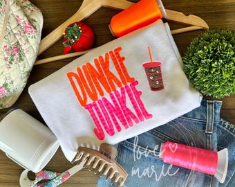 以下が含まれることがあります： ピンクとオレンジの文字で「Dunkie Junkie」と書かれた白いスウェットシャツ。スウェットシャツの右側に、茶色と白のアイスココアカップが刺繍されています。