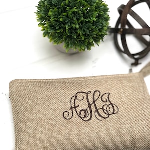 May include: A beige burlap zippered pouch with embroidered initials in a brown script font. A small green plant in a gray pot and a decorative metal sphere are in the background. The pouch is rectangular and has a simple design.