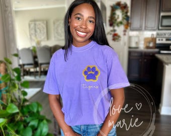 May include: A purple t-shirt with a gold paw print and the word "Tigers" printed on the front. The shirt is a relaxed fit and has a distressed look.