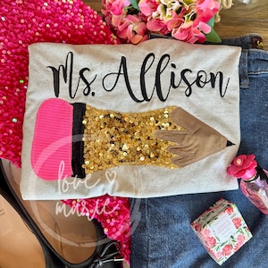 May include: A white t-shirt with the text "Ms. Allison" embroidered in black. A gold sequin pencil with a pink eraser is sewn onto the shirt.