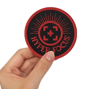Op de afbeelding: Een rood en zwart geborduurd patch met de woorden "HYPER FOCUS" en een kruisvormig symbool in het midden. De patch heeft een zonnestralenontwerp rond de rand.