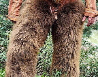 Puede incluir: Chaps de piel marrón con cinturón de cuero y detalles de cordones. Los chaps se llevan con una camisa abotonada estampada. La imagen está tomada al aire libre, con follaje verde al fondo.