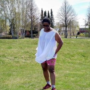 Puede incluir: Una persona con una blusa blanca y fluida y pantalones cortos estampados. Los pantalones cortos son rosas con un diseño circular repetitivo. La persona lleva gafas de sol y zapatillas granates, de pie sobre hierba con árboles al fondo.