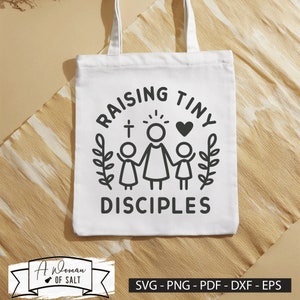 以下が含まれることがあります： 黒い文字で「Raising Tiny Disciples」と書かれた白いトートバッグ。女性と2人の子供、そして女性の頭上に十字架とハートのグラフィックが描かれている。