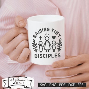 以下が含まれることがあります： 白いセラミック製のマグカップに、黒いデザインで「Raising Tiny Disciples」と書かれており、スタイリッシュなデザインで、母親と2人の子供が手をつないでいる様子が描かれています。母親は後光を放ち、頭上に十字架があります。デザインは葉の輪で囲まれています。