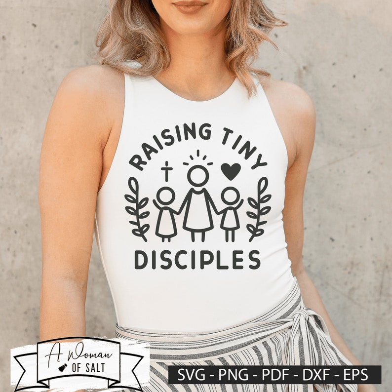 以下が含まれることがあります： 「Raising Tiny Disciples」と書いてある黒いグラフィックデザインが施された白いタンクトップ。グラフィックには、ハローと十字架をつけた女性と、手をつないでいる2人の子供が描かれています。
