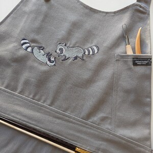 May include: Gray apron with embroidered raccoons, two brown buttons, and a pocket containing art tools. The apron has a waist tie and holds paint brushes. The apron is made of a durable fabric.