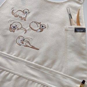 May include: A beige canvas apron with a large pocket on the right side. The pocket has a brown label with the text "CoCo Com Creations" on it. The apron has four embroidered images of otters in different poses. Two wooden tools are in the pocket and two paintbrushes are on the bottom of the apron.