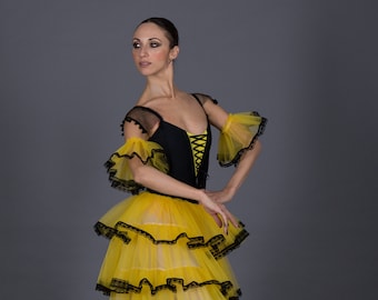 May include: A ballerina wearing a black and yellow layered tulle skirt with black lace trim and a black bodice with a lace-up front. She is wearing pointe shoes and has her hair pulled back.
