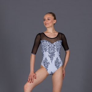 K&ouml;nnte beinhalten: Hellblaues Trikot mit schwarzem floralem Design und transparenten &Auml;rmeln. Das Trikot hat einen Sweetheart-Ausschnitt und wird mit Ballettschuhen getragen. Die T&auml;nzerin posiert vor grauem Hintergrund.